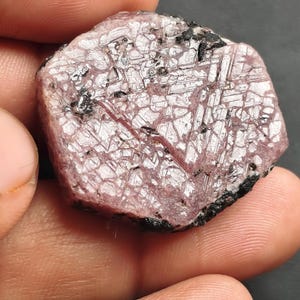 Puede incluir: Una gema rosa en bruto con una estructura cristalina e inclusiones oscuras. La piedra se sostiene en una mano sobre un fondo oscuro. La palabra "Photographer" está escrita en la parte inferior.