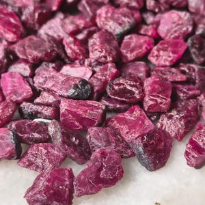 Puede incluir: Una colección de piedras preciosas de rubí en bruto y sin pulir en tonos profundos de magenta y rosa oscuro. Estos fragmentos minerales naturales presentan bordes irregulares y dentados, con una superficie cristalina y texturizada de opacidad variable.