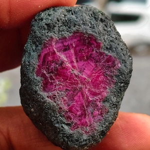 Puede incluir: Una piedra preciosa en bruto y sin pulir que presenta un centro cristalino de color magenta rosado intenso, encerrado en una matriz mineral exterior rugosa de color gris carbón oscuro a negro. El centro muestra un patrón estrellado.