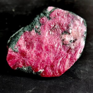 Puede incluir: Un espécimen mineral en bruto y sin pulir que presenta un centro cristalino de color magenta rosado intenso y vibrante, rodeado por una capa exterior de color verde bosque oscuro y texturizado. La superficie de la piedra muestra una mezcla de texturas rugosas, mates y reflectantes.