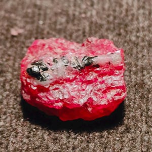 May include: A raw, vibrant red gemstone with a rough, crystalline texture. The stone features prominent inclusions of dark, metallic black mineral deposits and small patches of translucent white material embedded across its uneven surface.
