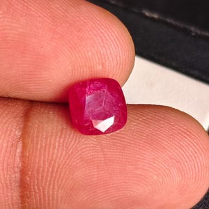 May include: A cushion-cut ruby gemstone with a vibrant, saturated red color and a faceted surface that reflects light. The gemstone displays internal inclusions and a clear, polished appearance, showcasing its deep red hue and geometric shape.