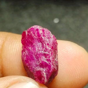 Puede incluir: Una piedra preciosa de rubí en bruto y sin pulir con un color rojo magenta profundo y vibrante. La piedra presenta una superficie cristalina naturalmente texturizada con facetas irregulares y secciones opacas, mostrando su forma mineral auténtica y sin tratar.