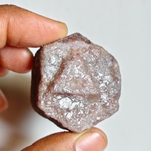 May include: A raw, hexagonal-shaped gemstone with a dusty, muted mauve and brownish-gray color palette. The surface of the stone displays a crystalline, textured appearance with a distinct triangular pattern etched into the center of the face.
