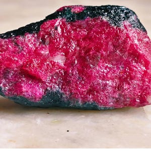Puede incluir: Un espécimen mineral en bruto y sin pulir que presenta un centro cristalino de color rosa magenta intenso y vibrante, encerrado en una corteza exterior de color negro mate oscuro. La piedra tiene una forma irregular y dentada.
