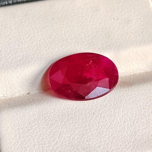 May include: A vibrant, oval-cut ruby gemstone with a deep red hue and multifaceted surface. The stone displays a polished, reflective finish with visible internal clarity and light refraction, showcasing a classic, symmetrical oval shape.