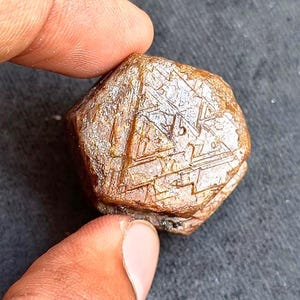 Puede incluir: Una gema marrón facetada con un patrón geométrico complejo. La piedra tiene una textura natural rugosa y se sostiene entre dos dedos sobre un fondo oscuro. La superficie de la gema refleja la luz, resaltando su intrincado diseño.