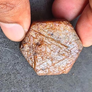 Puede incluir: Una gema facetada, hexagonal, de color marrón cálido. La superficie muestra intrincadas líneas que se cruzan, creando una apariencia texturizada. La gema se sostiene entre dos dedos, mostrando su tamaño y detalle.