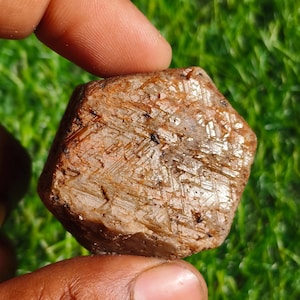 Könnte beinhalten: Ein grob geschnittener, sechseckiger Edelstein in Braun- und Beigetönen. Die Oberfläche hat ein strukturiertes Aussehen mit sichtbaren kristallinen Strukturen. Der Edelstein wird zwischen zwei Fingern vor einem Hintergrund aus grünem Gras gehalten.