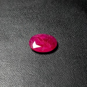 Può includere: Una gemma di rubino sfaccettata, di forma ovale. La gemma è di un rosso intenso e vibrante, con una sfaccettatura bianca brillante che riflette la luce. Il rubino è posto su uno sfondo nero strutturato.