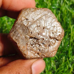 Puede incluir: Una gema en bruto de forma hexagonal, de color marrón claro y con una superficie texturizada. La piedra presenta una estructura cristalina con líneas y patrones visibles. La gema se sostiene en una mano sobre un fondo de hierba verde.