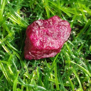 Peut inclure: Une pierre précieuse brute et non polie d'une couleur magenta profond et rouge foncé. La pierre présente une forme irrégulière et dentelée avec une surface cristalline texturée qui reflète la lumière sur ses facettes inégales.