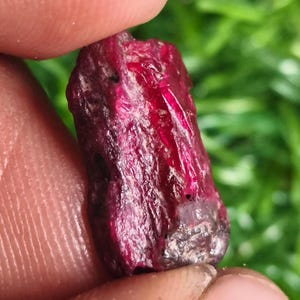 Puede incluir: Una piedra preciosa de rubí en bruto y sin tallar con un color rojo intenso y una superficie cristalina texturizada. La piedra presenta secciones translúcidas que captan la luz, mostrando variaciones naturales en su estructura.