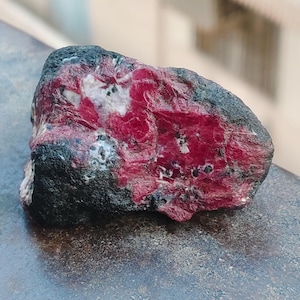 May include: A raw, unpolished mineral specimen featuring a vibrant, deep red crystalline center contrasted by a rough, dark gray to black outer crust. The stone displays irregular, jagged edges and a textured surface with small white and black inclusions.