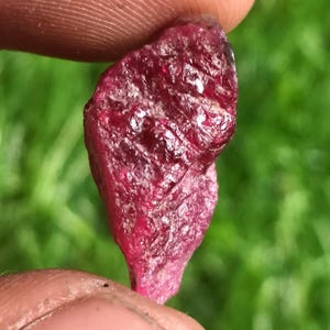 Peut inclure: Une pierre précieuse rubis brute et non polie d'une couleur rouge profond et saturée. La pierre présente une surface naturellement irrégulière et texturée avec des facettes cristallines visibles et une qualité translucide qui capte la lumière.