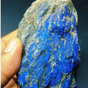 Pode incluir: Um pedaço bruto e não polido de mineral lápis-lazúli com uma cor azul profunda e vibrante, apresentando inclusões naturais cinzentas e brancas visíveis por toda a textura de superfície cristalina e irregular do espécime.