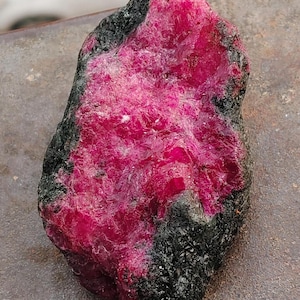 Könnte beinhalten: Ein rohes, unpoliertes Mineralexemplar mit einem leuchtenden, tief magentafarbenen, kristallinen Zentrum, das von einer dunklen, strukturierten, kohleschwarzen äußeren Matrix umgeben ist. Die Oberfläche des Steins zeigt einen Kontrast zwischen den rauen, dunklen Kanten und der funkelnden, gezackten, kristallinen Struktur des rosa Kerns.