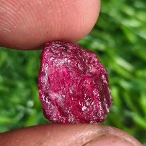 Peut inclure: Une pierre précieuse rubis brute et non taillée, d'une couleur rouge profond et saturée, avec une surface naturellement texturée et à facettes. La pierre présente une qualité translucide avec des inclusions internes visibles et des bords irréguliers.