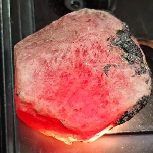 Puede incluir: Una gema natural en bruto, de color rosa rojizo. La piedra tiene una superficie texturizada con zonas de color gris oscuro y negro. La parte inferior de la piedra brilla con una luz roja brillante, creando un contraste llamativo.