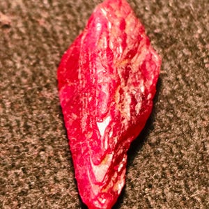 May include: A raw, elongated crystal specimen with a deep, vibrant red colour. The surface of the stone features a complex, jagged texture with natural facets and ridges that catch the light, creating varied highlights across its irregular shape.