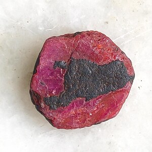 May include: A rough, uncut ruby gemstone with a deep red hue and a dark, charcoal-colored inclusion. The stone's surface is textured, and it sits against a white background.