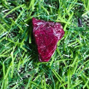 Peut inclure: Une pierre précieuse brute et non polie d'une couleur rouge magenta profonde et saturée. La pierre présente une forme irrégulière et anguleuse avec une texture cristalline à facettes qui reflète la lumière sur sa surface inégale.