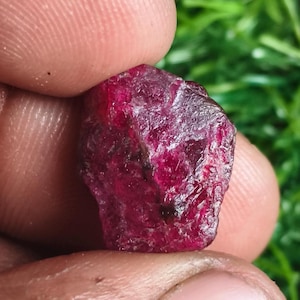 May include: A raw, unpolished gemstone with a deep, saturated magenta-red color. The stone features an irregular, multifaceted surface with natural textures, visible crystalline structures, and translucent areas that catch the light.