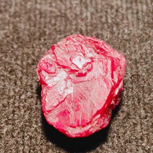 Puede incluir: Una piedra preciosa en bruto y sin pulir de un color rojo intenso y vibrante. La piedra presenta una forma irregular y dentada con múltiples facetas y texturas superficiales naturales, mostrando una estructura cristalina.