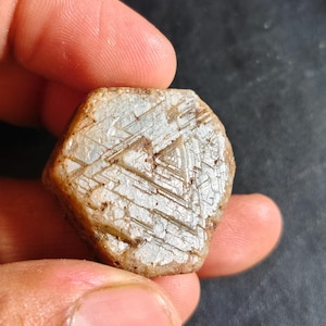 Può includere: Una gemma grezza, di colore chiaro, con un motivo geometrico unico. La pietra ha un aspetto naturale e non lucido, con una miscela di tonalità beige e bianco. La superficie presenta una serie di linee e forme che si intersecano, creando un effetto strutturato.
