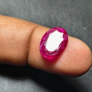 May include: A faceted, oval ruby gemstone. The gem is a vibrant, deep pinkish-red colour with internal inclusions. The cut of the stone allows light to reflect, creating a bright, sparkling appearance. The ruby is displayed against a neutral background.