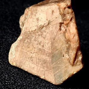 May include: A roughly-shaped, light brown gemstone with a faceted surface. The stone has a natural, earthy appearance with subtle variations in colour and texture. The gemstone is displayed against a dark background.