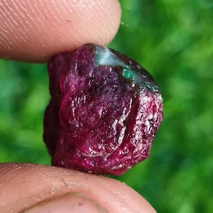 Può includere: Una gemma di rubino grezza, non tagliata, tenuta tra le dita. Il rubino è di un rosso intenso con aree verdi e bianche. La superficie ha un aspetto strutturato e naturale, che mostra la sua forma grezza. Lo sfondo è verde sfocato.
