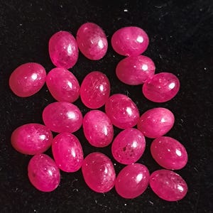 May include: A collection of fifteen small, oval-shaped, vibrant pink gemstones arranged in a cluster. Each stone features a smooth, polished cabochon surface that reflects light, showcasing a consistent deep magenta hue across the entire set.