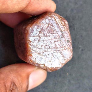 Pode incluir: Uma gema bruta, castanho-avermelhada, com uma superfície facetada. A pedra tem um padrão geométrico de linhas e ângulos. A gema é segurada entre os dedos. Parece ser um espécime mineral natural.