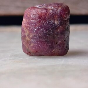 May include: A raw, cylindrical ruby gemstone with a deep magenta and dark red hue. The stone features a textured, uneven surface with natural striations and a matte finish, displaying a solid, opaque appearance without any visible facets.