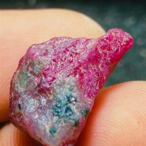 May include: A rough-cut ruby gemstone with a deep red hue. The surface has a textured appearance with areas of lighter pink and patches of blue-green mineral inclusions. The gemstone is held in a person's hand.