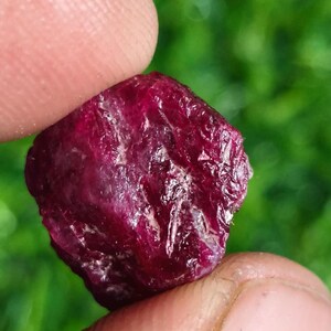 May include: A raw, deep red ruby gemstone with a textured, multifaceted surface. The stone displays natural inclusions and a translucent quality, showing variations in colour intensity across its irregular, organic shape and crystalline structure.
