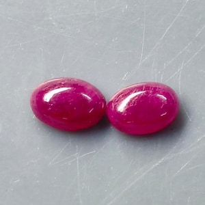Puede incluir: Dos piedras preciosas de forma ovalada, talladas en cabujón, con un color magenta rosado intenso y vibrante. Cada piedra presenta una superficie lisa y pulida con un reflejo de luz blanco, resaltando el perfil redondeado de las gemas.
