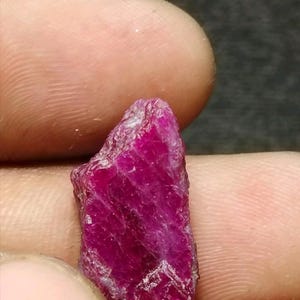 May include: A raw, unpolished ruby gemstone with a deep, vibrant magenta-red colour. The stone features a jagged, irregular crystalline structure with natural facets and a translucent quality that catches the light across its textured surface.