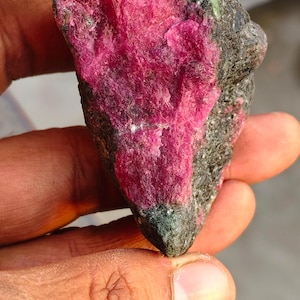 May include: A raw, unpolished mineral specimen featuring a vibrant, deep pink crystalline section embedded within a dark, textured, greyish-green rock matrix. The stone has an irregular, angular shape with a rough, natural surface texture.