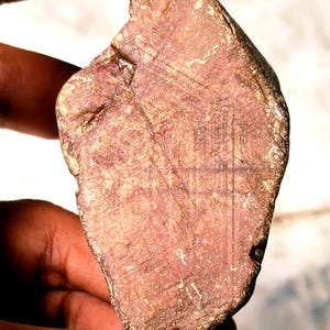 May include: A roughly-cut, reddish-brown gemstone with a textured surface. The stone has a natural, irregular shape and is held between two fingers. The surface shows subtle variations in colour and texture, with visible lines.