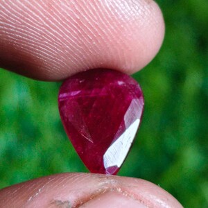 May include: A pear-shaped, deep red gemstone with a faceted surface. The stone exhibits internal inclusions and a polished finish, reflecting light on its angled facets. The gem is held between two fingers, showcasing its vibrant, saturated color.