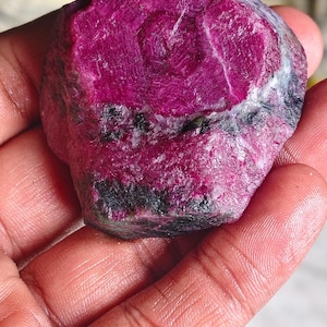 May include: A raw, unpolished ruby gemstone with a deep magenta and crimson hue. The stone features a hexagonal crystalline structure on its top surface, with visible dark mineral inclusions and rough, textured edges throughout its form.