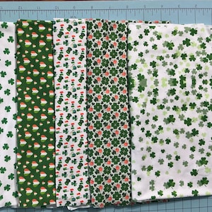 May include: A collection of folded fabric pieces featuring St. Patrick's Day patterns. The fabrics display various designs, including green shamrocks on white, and patterns with leprechaun hats and orange accents. The fabrics are arranged on a blue cutting mat.