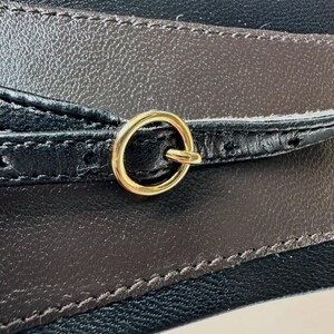 Peut inclure: Gros plan sur une ceinture en cuir noir et marron avec une boucle en anneau en m&eacute;tal dor&eacute;. La ceinture pr&eacute;sente un design cousu et une surface textur&eacute;e. La boucle est circulaire et fix&eacute;e &agrave; la ceinture.