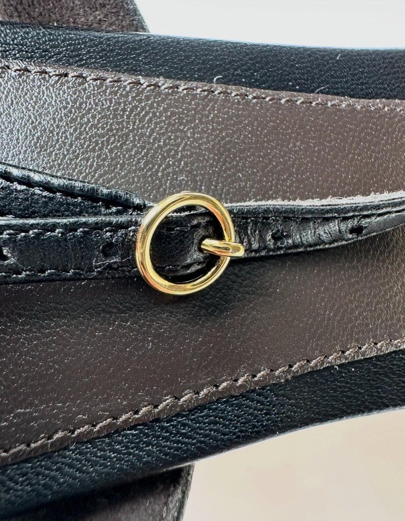 Peut inclure: Gros plan sur une ceinture en cuir noir et marron avec une boucle en anneau en m&eacute;tal dor&eacute;. La ceinture pr&eacute;sente un design cousu et une surface textur&eacute;e. La boucle est circulaire et fix&eacute;e &agrave; la ceinture.