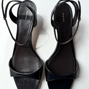 Peut inclure: Paire de sandales en cuir noir avec fines lani&egrave;res de cheville. Les sandales ont un design minimaliste avec un bout arrondi et le nom de la marque "THE ROW" imprim&eacute; sur la semelle int&eacute;rieure. Fond blanc.