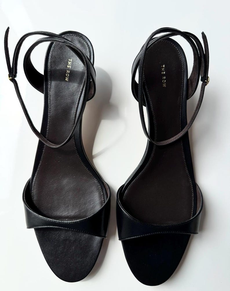 Peut inclure: Paire de sandales en cuir noir avec fines lani&egrave;res de cheville. Les sandales ont un design minimaliste avec un bout arrondi et le nom de la marque "THE ROW" imprim&eacute; sur la semelle int&eacute;rieure. Fond blanc.