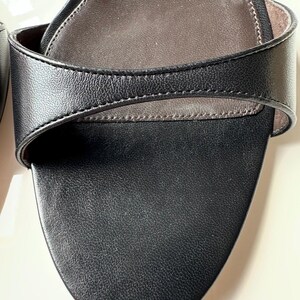 Peut inclure: Gros plan d'une sandale en cuir noir. La sandale pr&eacute;sente un bout arrondi et une bride qui se courbe sur le pied. La semelle int&eacute;rieure est de couleur marron clair et les coutures sont visibles sur les bords des brides.