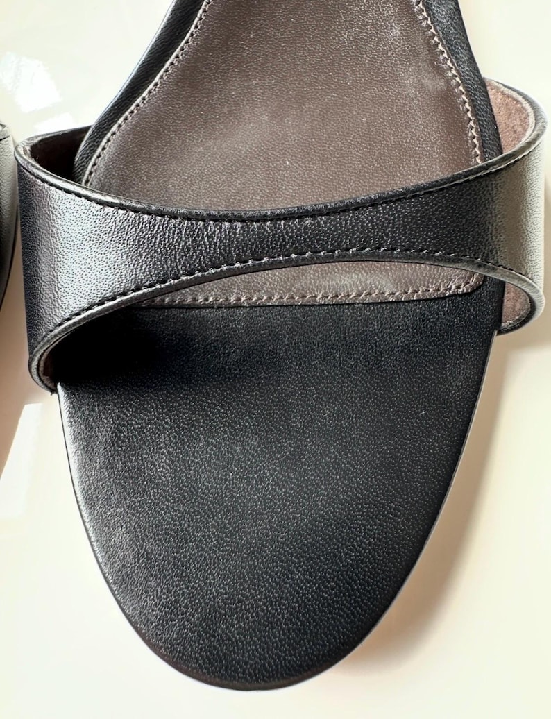 Peut inclure: Gros plan d'une sandale en cuir noir. La sandale pr&eacute;sente un bout arrondi et une bride qui se courbe sur le pied. La semelle int&eacute;rieure est de couleur marron clair et les coutures sont visibles sur les bords des brides.